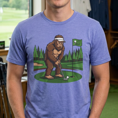 A man wearing a heathered periwinkle blue t-shirt with a cartoon graphic of a Bigfoot golfing, standing inside a golf shop.