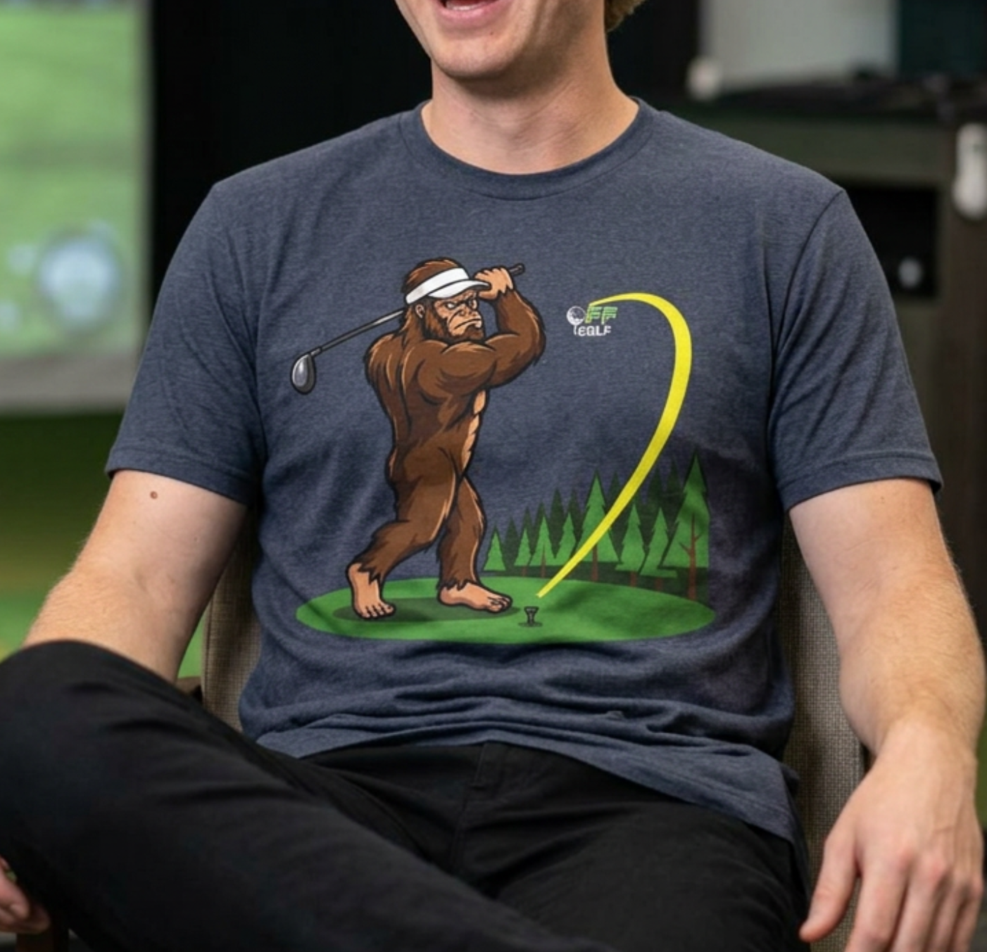 A man sitting in a chair wearing a dark heather gray t-shirt with a cartoon graphic of a bigfoot golf swing. The background shows a golf simulator screen and a desk setup.