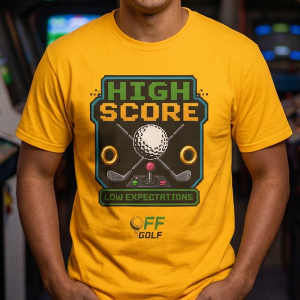 A smiling man wearing a bright yellow graphic t-shirt in an arcade room. The shirt features a retro-style green and black graphic that reads 'HIGH SCORE' with a golf ball and clubs, and 'OW EXPECTATIONS' below it, with 'OFF GOLF' printed underneath.