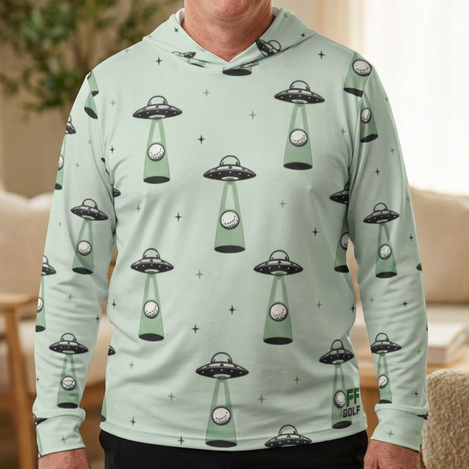 A smiling man wearing a light mint green long-sleeve hoodie with an all-over UFO pattern, standing in a cozy, sunlit living room.