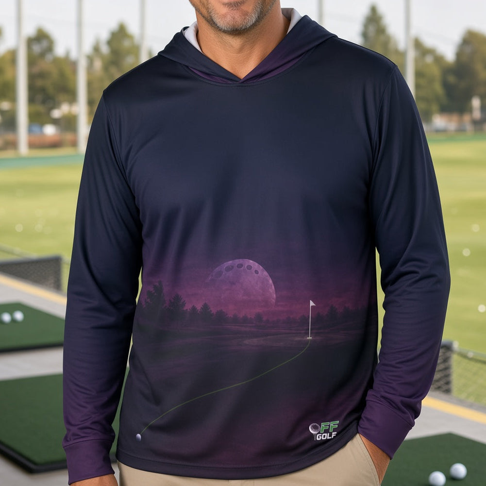 A man wearing a dark navy long-sleeve hoodie with a purple and pink moon landscape graphic on the front, standing on a golf driving range.