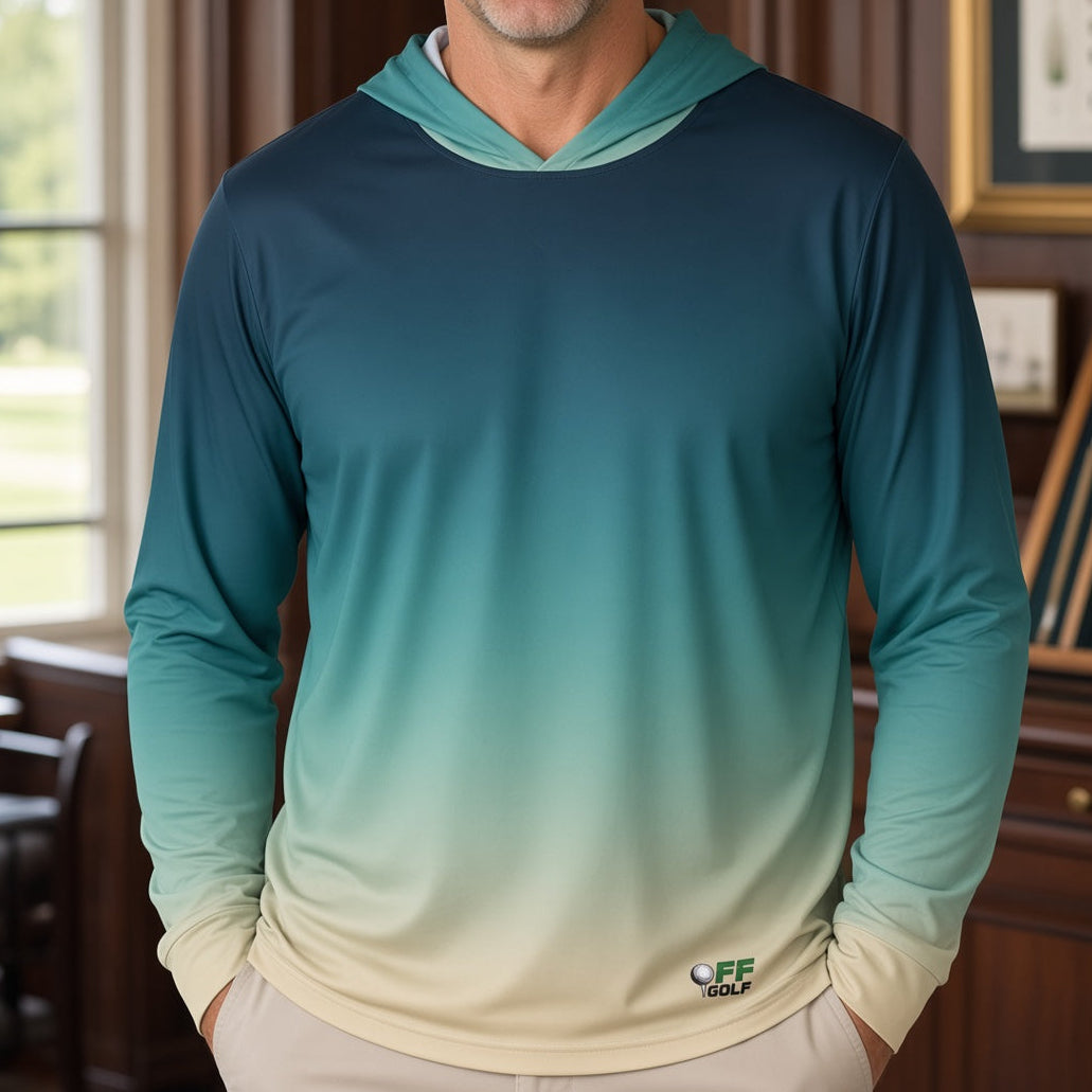 A smiling man stands indoors wearing a long-sleeve gradient hooded shirt that fades from dark teal at the top to light aqua and cream at the bottom. The room has wood paneling, a window, and framed items on a cabinet behind him.