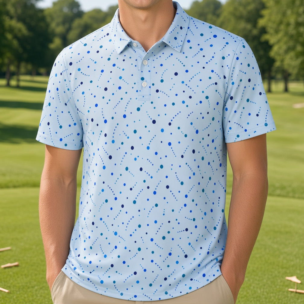 A smiling young man stands on a golf course wearing a light blue short-sleeve polo shirt with a blue and dark blue dotted pattern, paired with khaki pants.