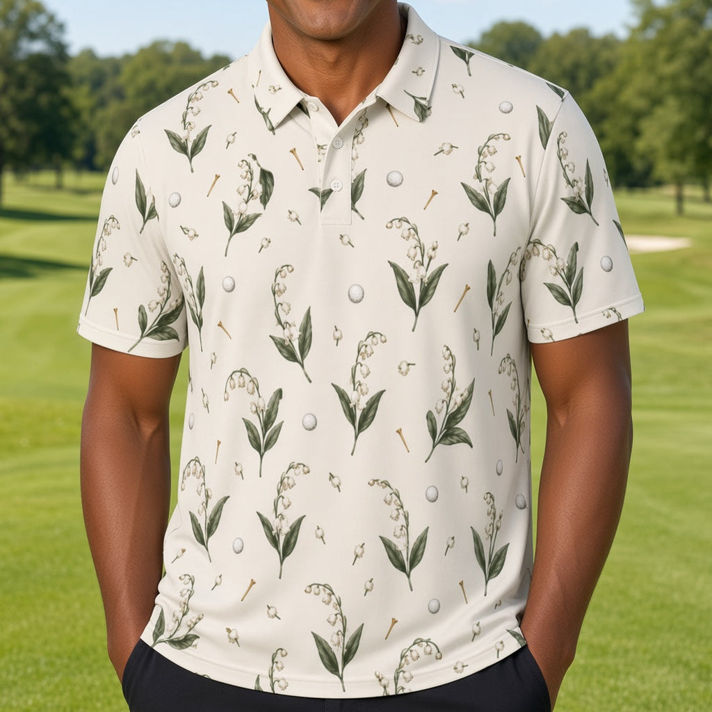 A smiling man wearing a short-sleeve polo shirt with a golf-themed botanical print stands on a lush green golf course. The shirt has an off-white base with green leaves, golf tees, and golf balls printed all over it.