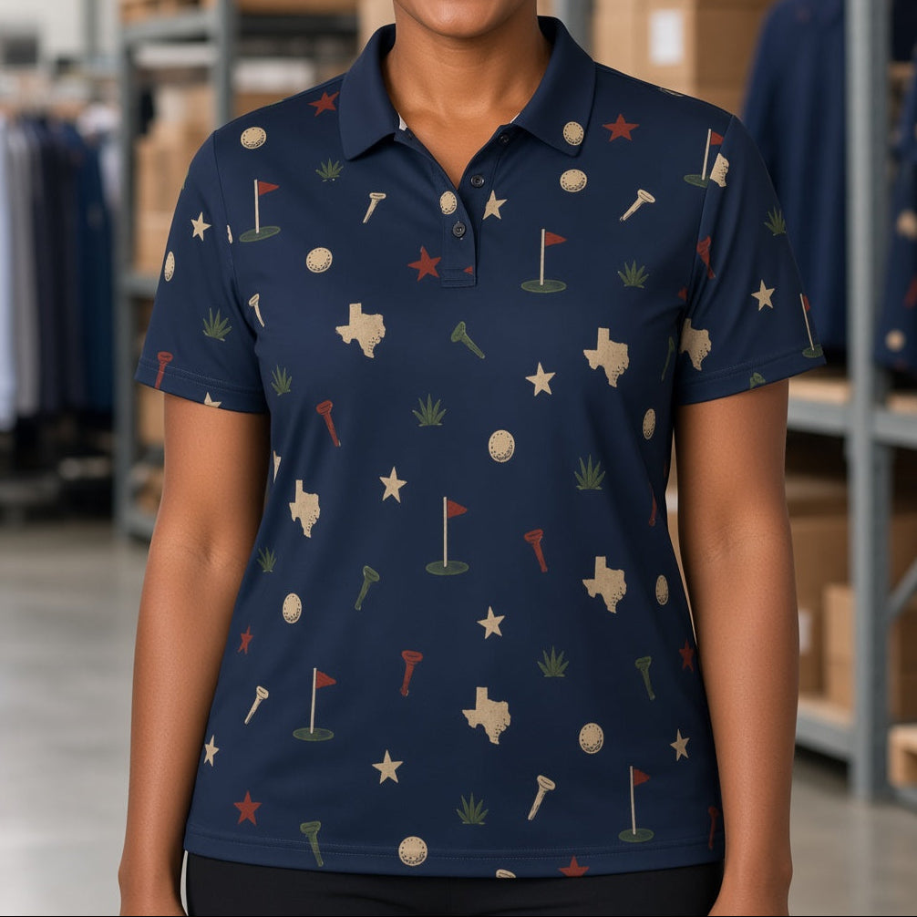A smiling woman with curly hair stands in a warehouse setting wearing a navy blue short-sleeve polo shirt with a golf-themed print of flags, tees, golf balls, and the state of Texas. Shelves with boxes and other clothing are visible in the background.