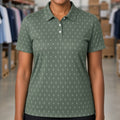 A smiling woman with short curly black hair stands in a warehouse setting wearing a sage green short-sleeve polo shirt with a small white repeating pattern. The background shows shelves stocked with cardboard boxes and folded clothing.