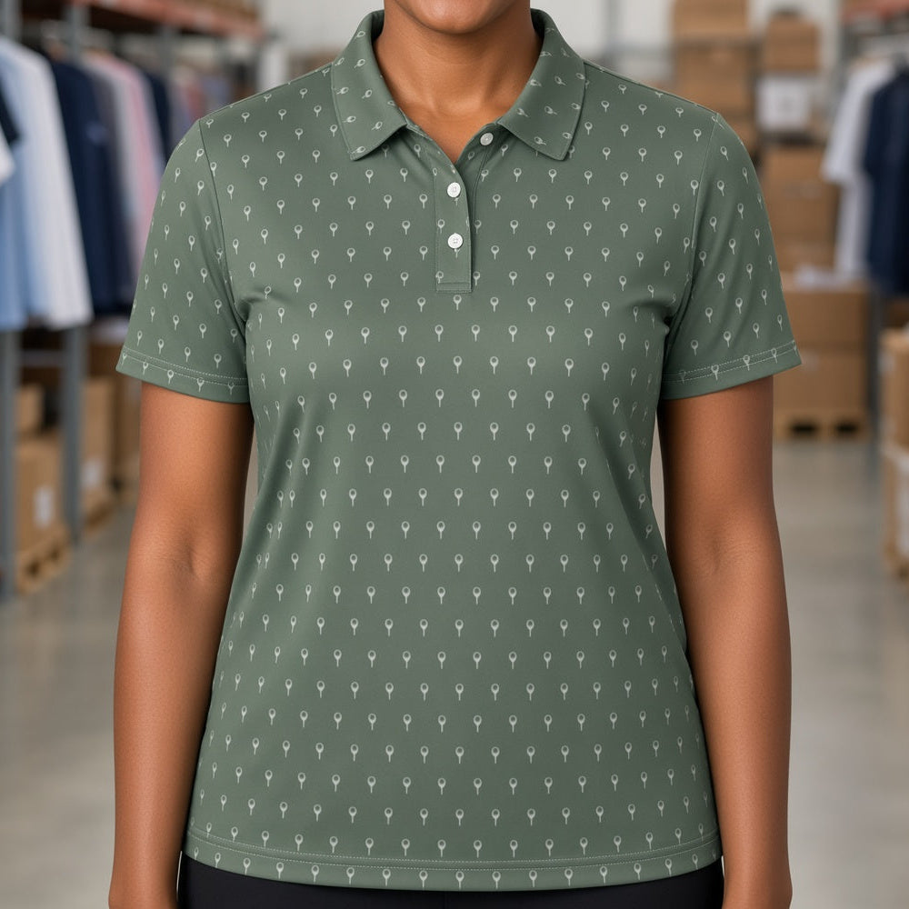 A smiling woman with short curly black hair stands in a warehouse setting wearing a sage green short-sleeve polo shirt with a small white repeating pattern. The background shows shelves stocked with cardboard boxes and folded clothing.