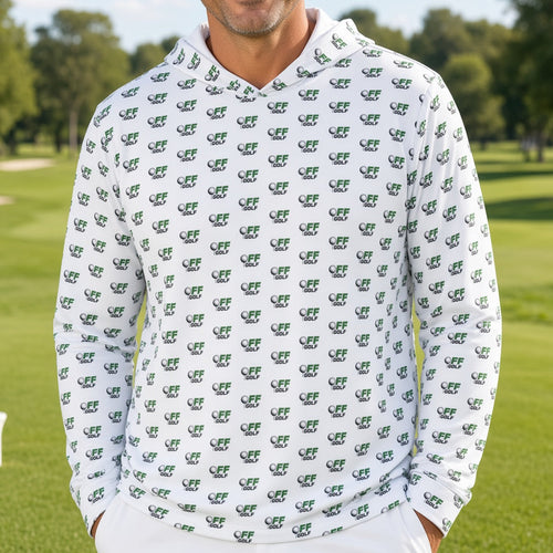 A smiling man stands on a golf course wearing a white long-sleeve hoodie covered in a repeating green crocodile pattern, paired with white pants.