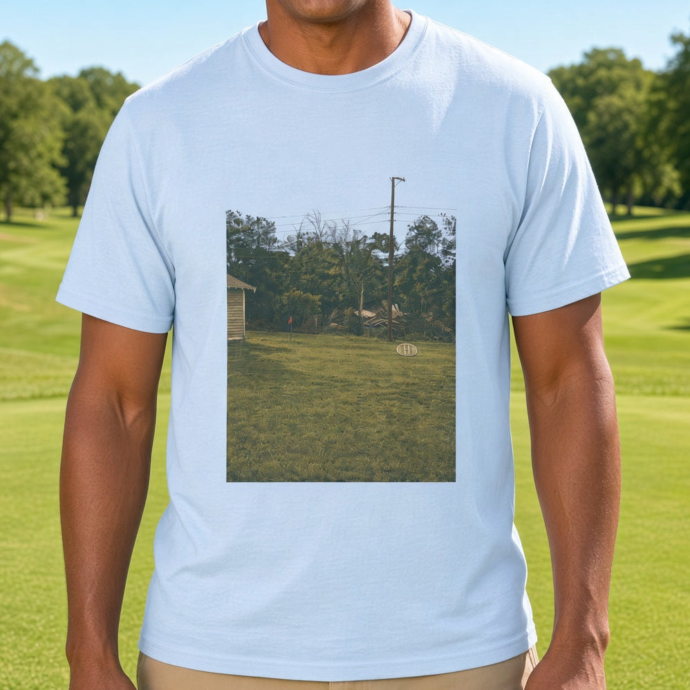 A smiling man wearing a light blue crewneck t-shirt with a rectangular photographic print on the front, standing on a sunny green golf course.