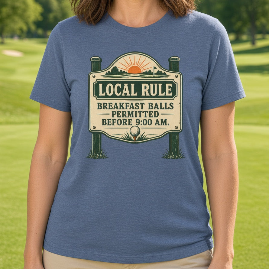 A woman stands outdoors on a golf course wearing a slate blue graphic t-shirt with a vintage-style sign that reads 'LOCAL RULE BREAKFAST BALLS PERMITTED BEFORE 5:00 AM'.