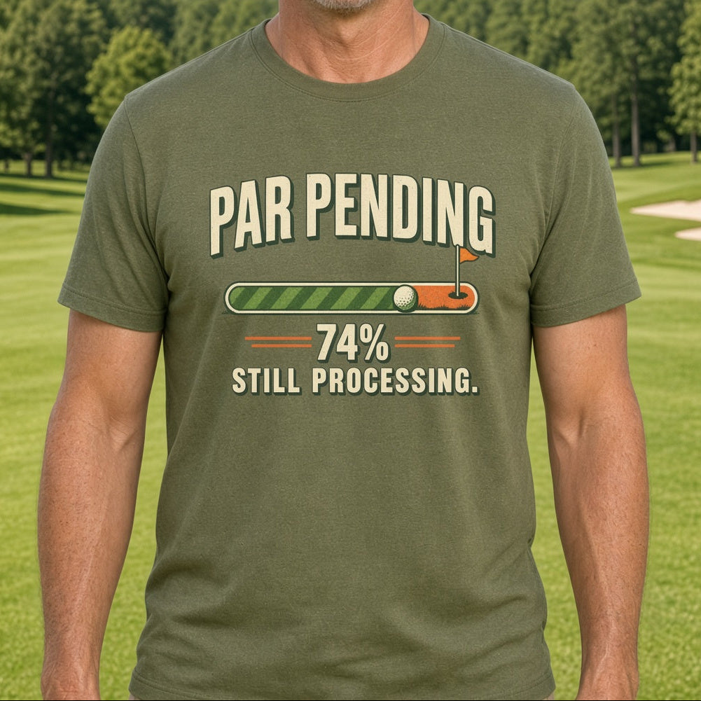 A smiling man wearing an olive green graphic t-shirt stands on a golf course. The t-shirt features a humorous graphic with a golf ball and flag on a progress bar that reads 'PAR PENDING' and '74% STILL PROCESSING.