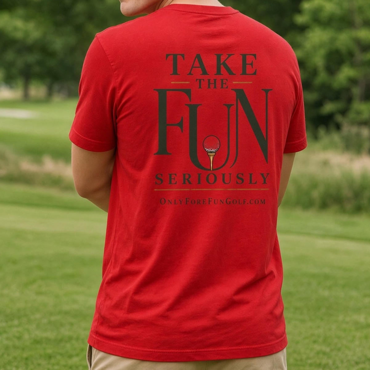 A man standing outdoors on a grassy field, shown from the back, wearing a bright red t-shirt with a dark green graphic text that reads 'TAKE THE FUN SERIOUSLY' and khaki pants.
