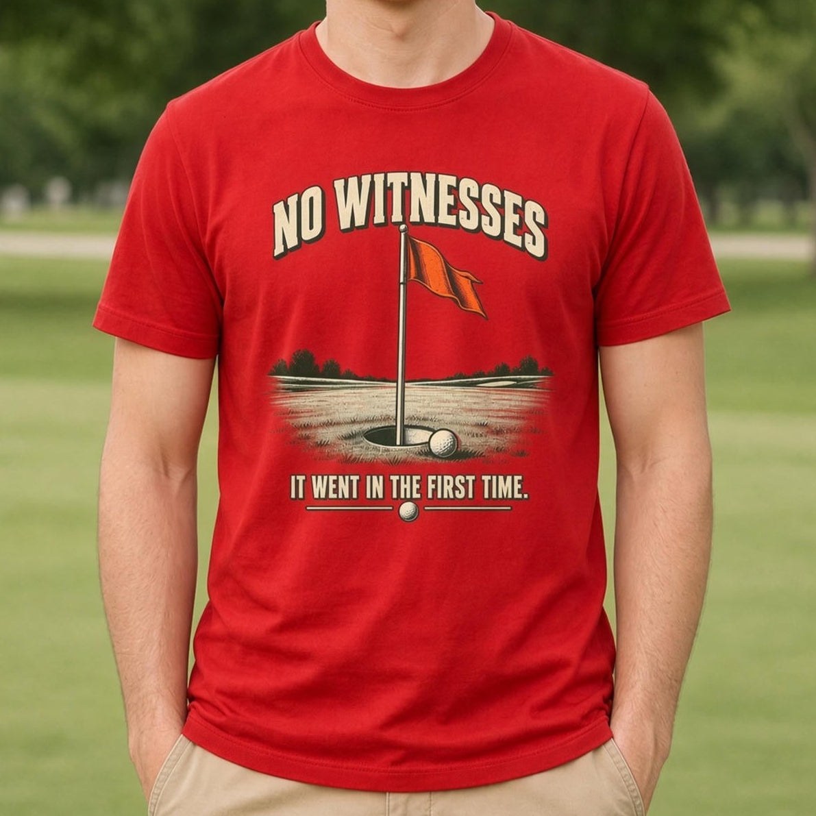 A smiling young man stands on a blurred golf course wearing a bright red graphic t-shirt with a golf hole illustration and the text 'NO WITNESSES IT WENT IN THE FIRST TIME', paired with khaki pants.