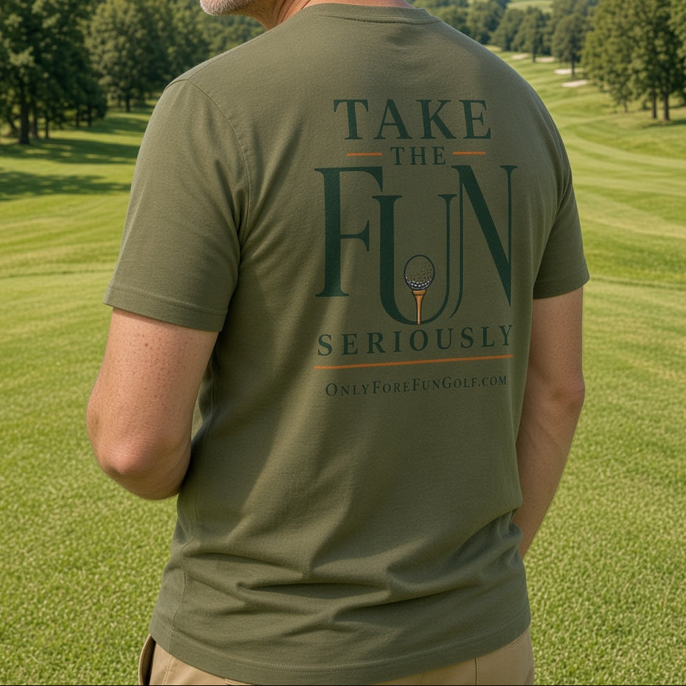 A man wearing an olive green t-shirt with a golf-themed graphic on the back, standing on a sunny golf course with trees and blue sky in the background.