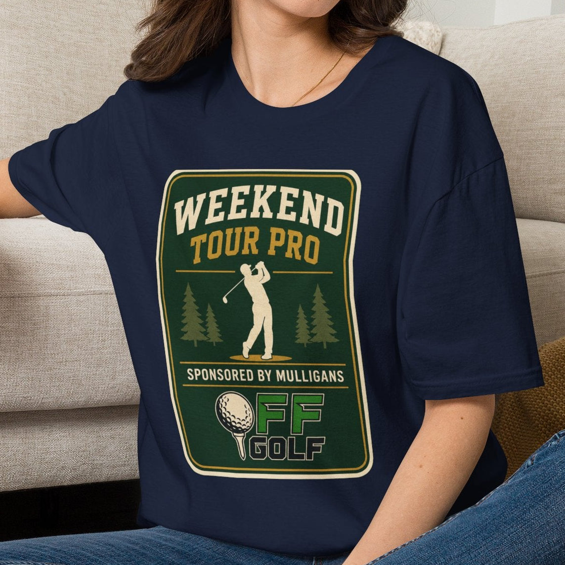 A woman sitting cross-legged on a woven rug in front of a beige sofa, wearing a navy blue graphic T-shirt with a golf-themed 'Weekend Tour Pro' print, blue jeans, and white sneakers.