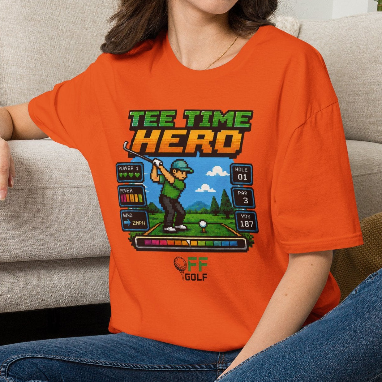 A woman sitting cross-legged on a woven rug in front of a beige sofa, wearing an orange graphic T-shirt with a retro golf machine design and the words 'TEE TIME HERO' and 'GOLF'.