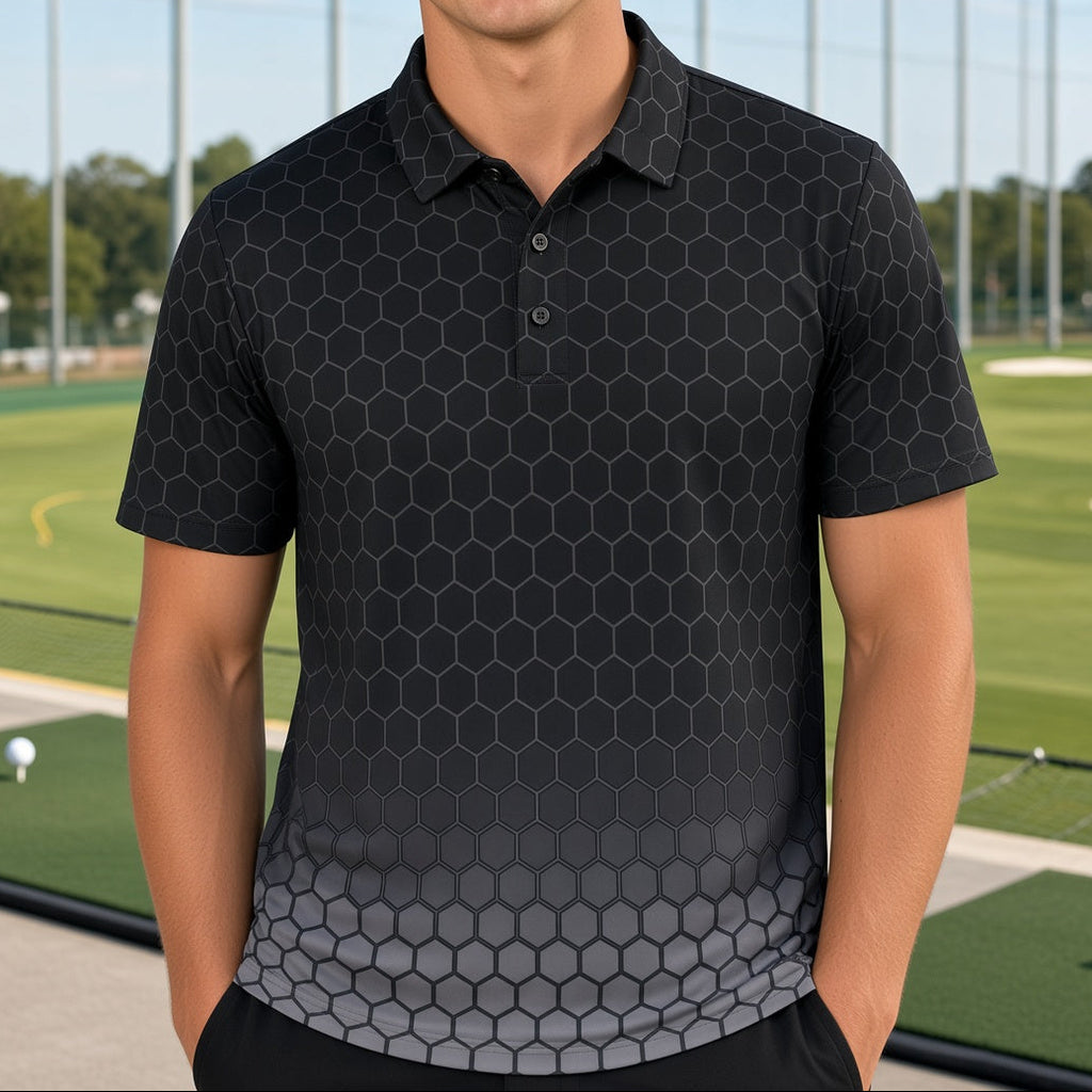 A smiling young man stands on a golf driving range wearing a black short-sleeve polo shirt with a subtle dark gray hexagonal pattern that fades into gray at the hem, against a background of green golf turf and netting.